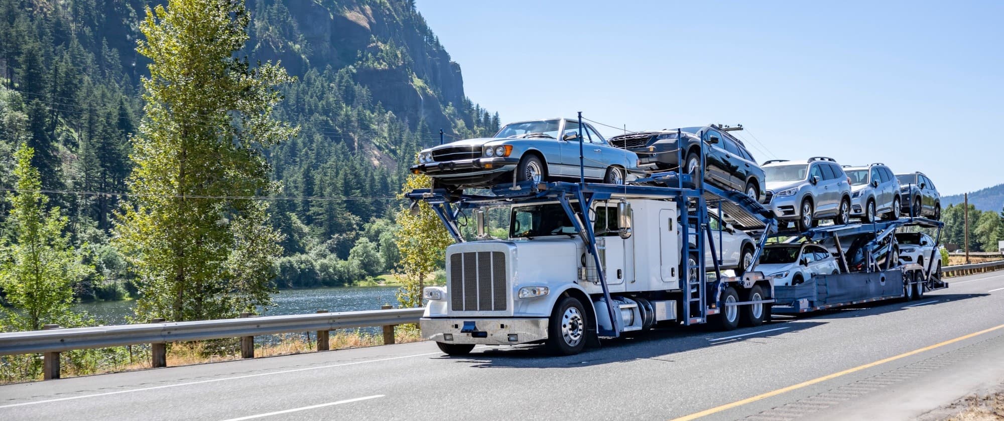White car carrier truck transporting vehicles on a scenic highway with a forested mountain backdrop.