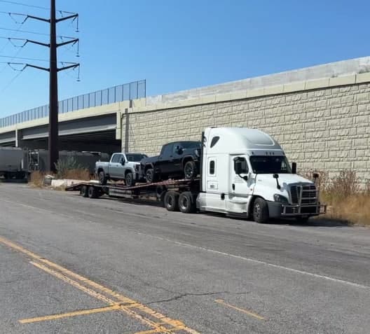 White semi-truck transporting two pickup trucks on a flatbed trailer parked beside a concrete wall.